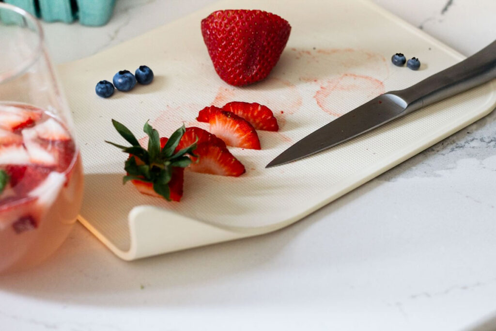 Cut strawberries on a cutting board with blueberries and a cocktail to the left.