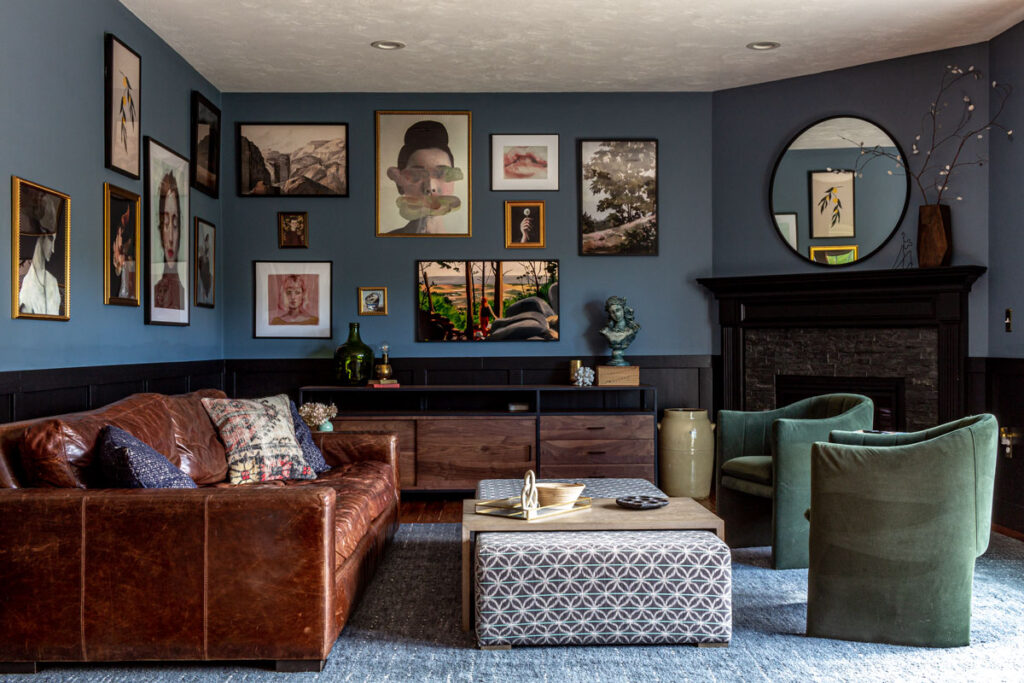 Blue and black living room with wainscoting board and batten, corner fireplace, and large gallery wall. Leather couch and green velvet chairs in a seating area.