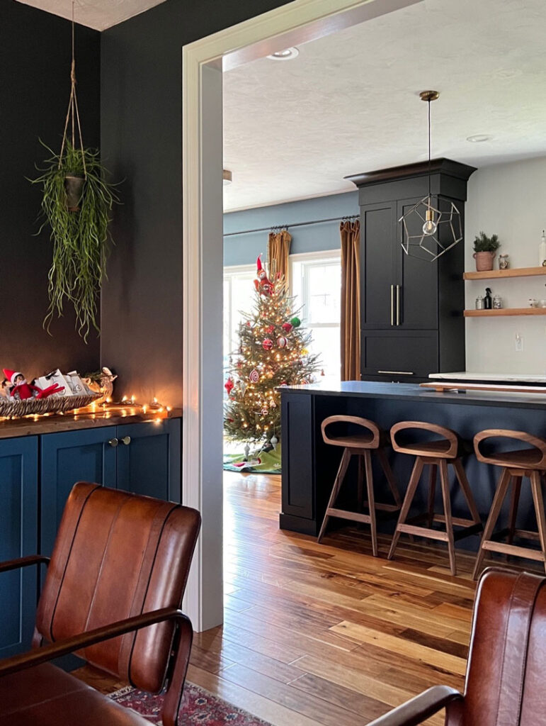 Black kitchen island with black kitchen cabinets, a Christmas tree in the background, geometric pendant lights and walnut counter stools. Leather chair and blue cabinets in the foreground.