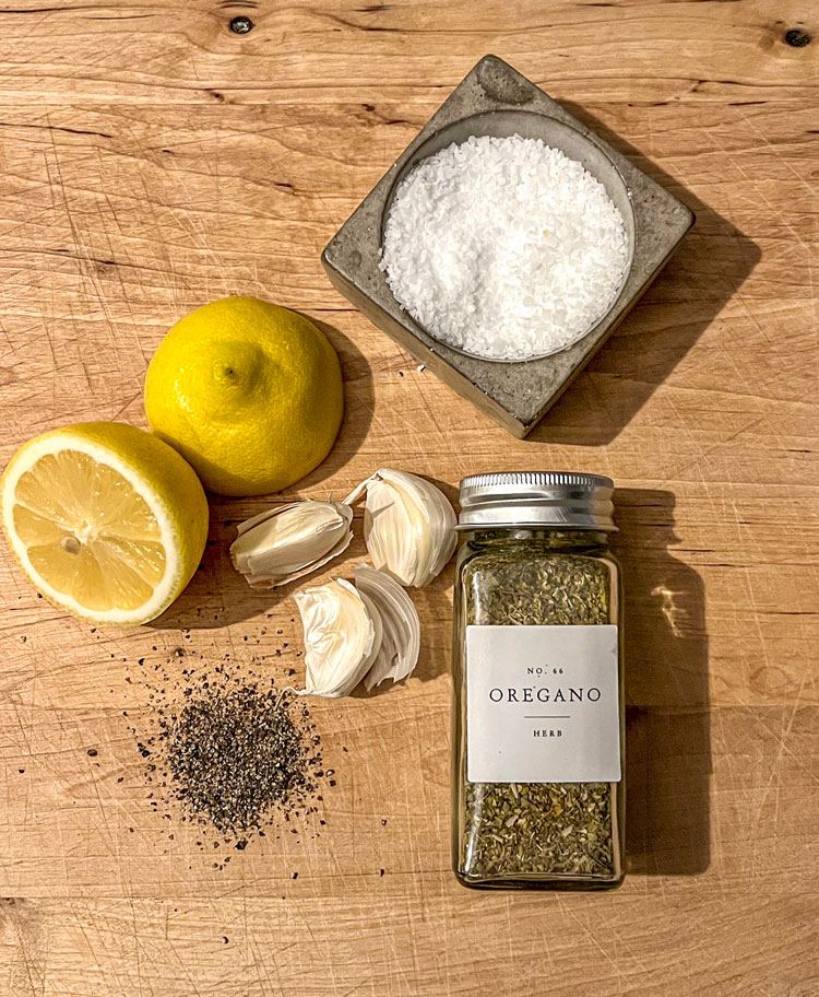 The ingredients for the perfect lemon garlic chicken lying on a wooden cutting board: fresh lemon, garlic cloves, salt, pepper, and oregano.