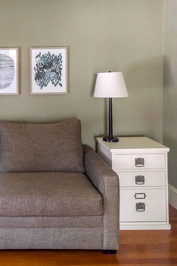 Brown couch in a green gray living room with abstract blue art on the walls and a table lamp on the white side table.
