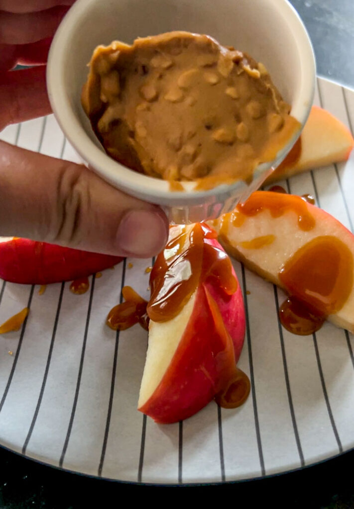 Hand holding a small white dish full of chunky peanut butter. Apple slices covered in caramel sauce on a plate in the background.