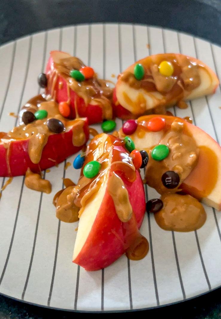 Sliced apples covered in peanut butter, mini M%Ms and caramel arranged on a plate.