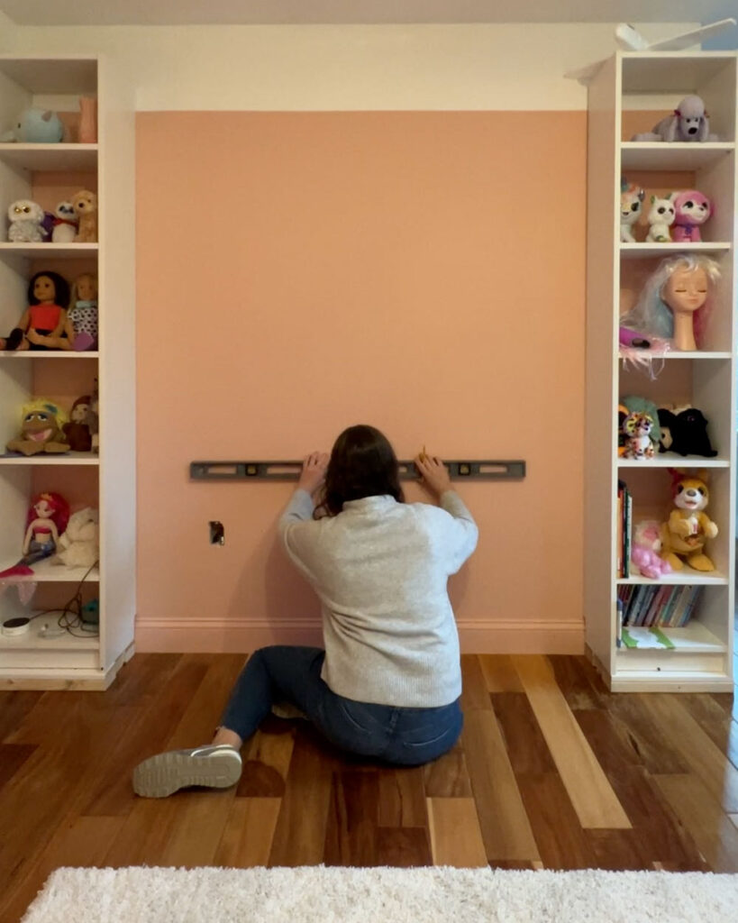 Woman holding a level against a pink wall between two bookcases.