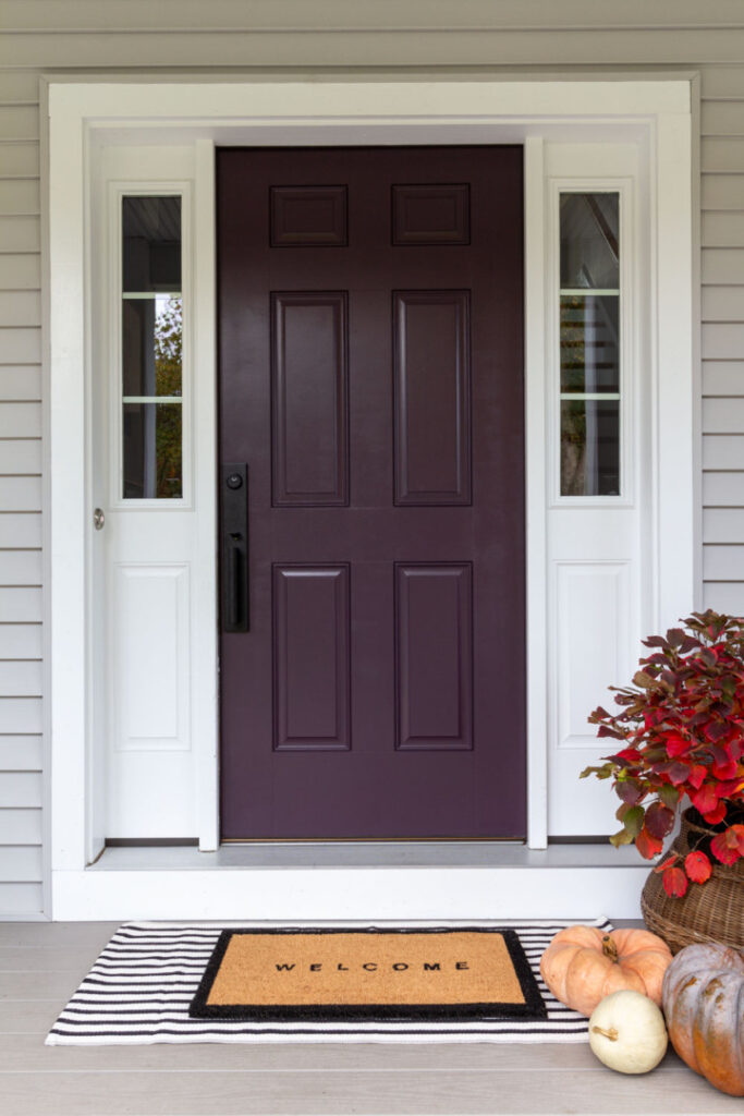 Purple fron door on a grey house. Striped welcome mat on the porch next to a red shrub and three pumpkins.
