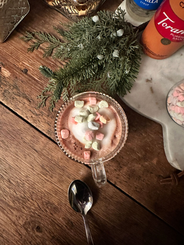 Cup of cocoa with colorful marshmallows sitting on a reclaimed wood counter decorated for Christmas.