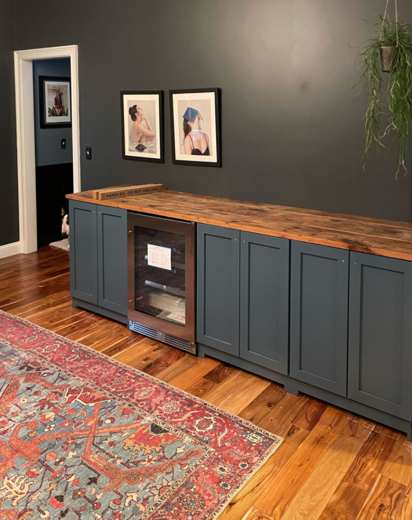 Blue cabinets with a mini fridge and reclaimed wood counter against a black wall with a vintage rug in the foreground. 